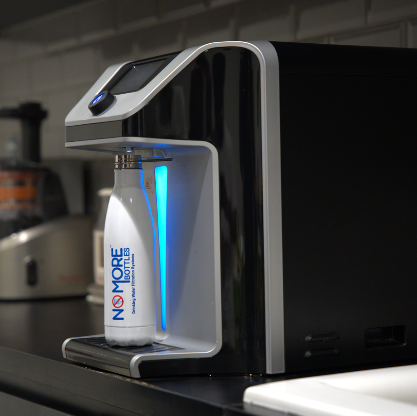 A countertop water filtration machine in a kitchen setting, dispensing a white bottle labeled 'NO MORE BOTTLES' under a blue light. 
